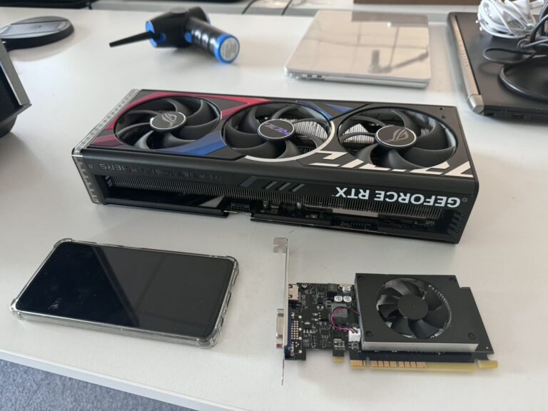 A large ASUS ROG GeForce RTX graphics card next to a small low-profile graphics card, a phone and an electric air duster on a workshop table